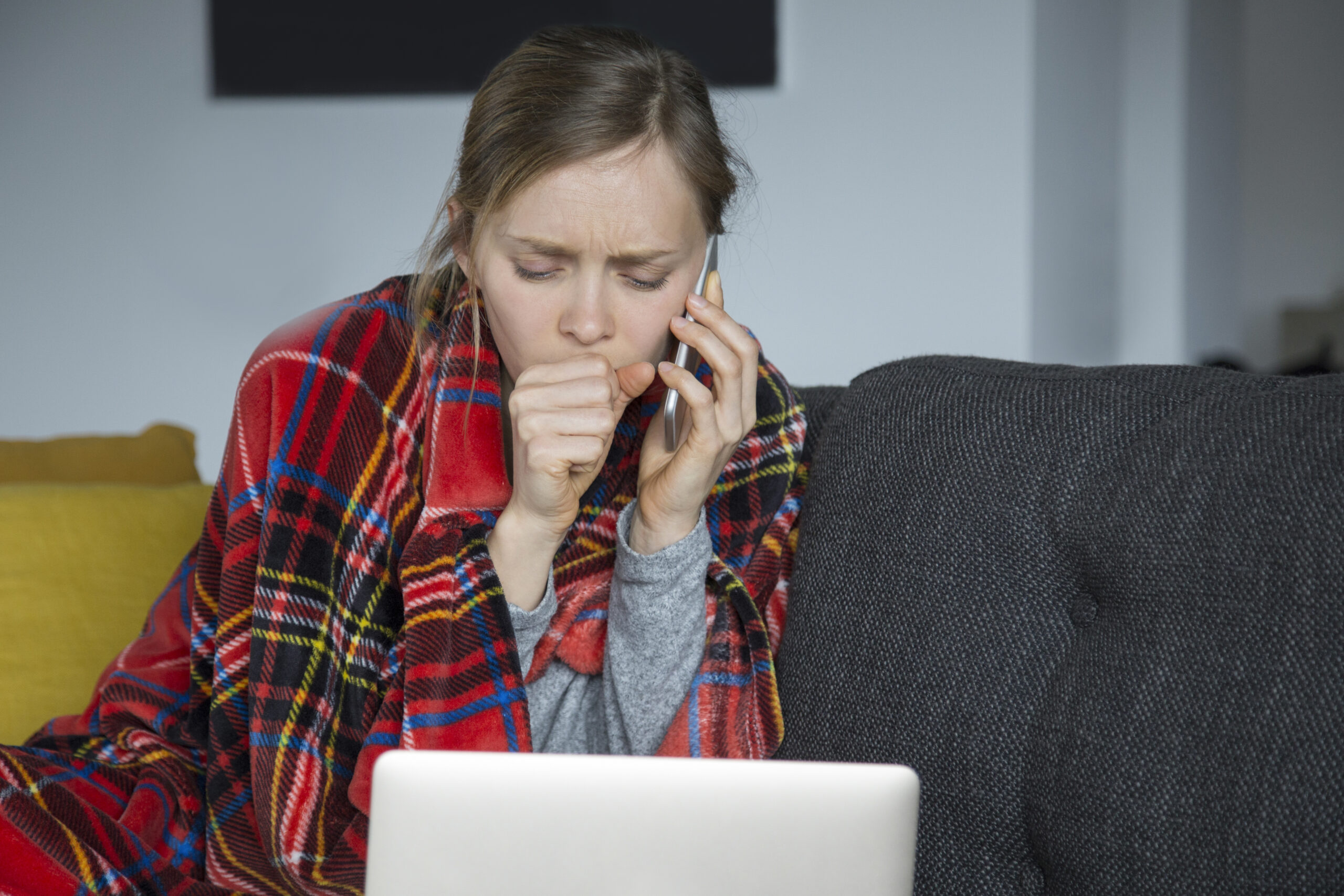 Sick young fair-haired Caucasian woman covered with plaid sitting on grey sofa in living room, coughing, working on laptop. Illness, work concept